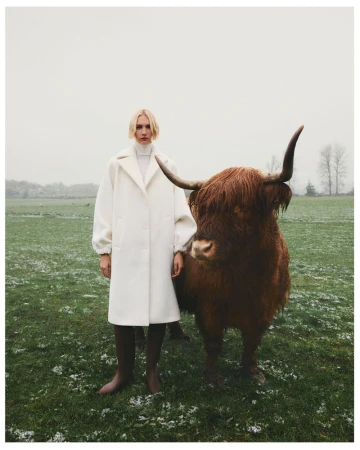 A blond model in a cream coat poses with a highland cow in a snowy, grassy field on an overcast day. Fashion editorial.