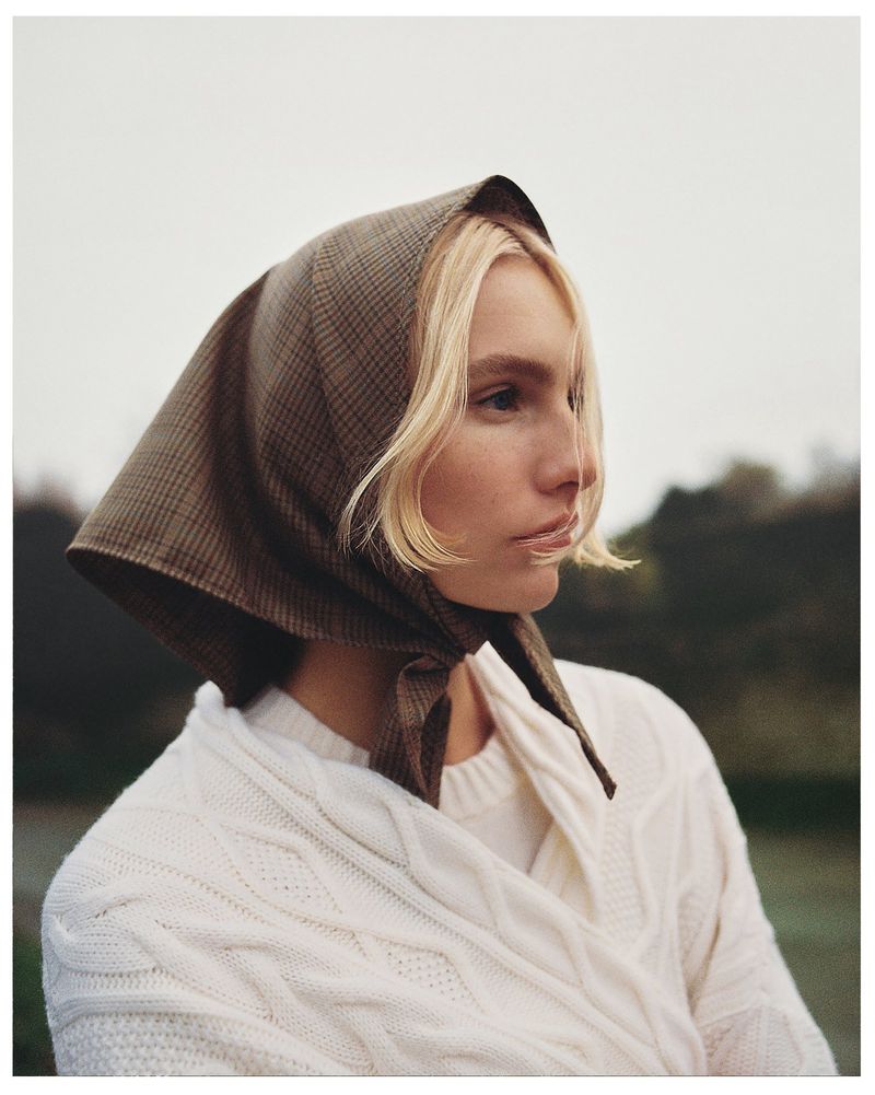 Portrait of a blonde woman wearing a brown patterned headscarf and cream-colored sweater, posing outdoors with trees and sky in the background.
