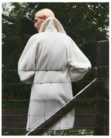 Model wearing a white wool coat standing outdoors near a wooden post and barbed wire fence.