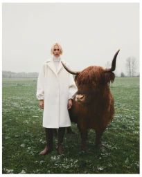 Model in white coat standing next to a highland cattle in a grassy field under an overcast sky.