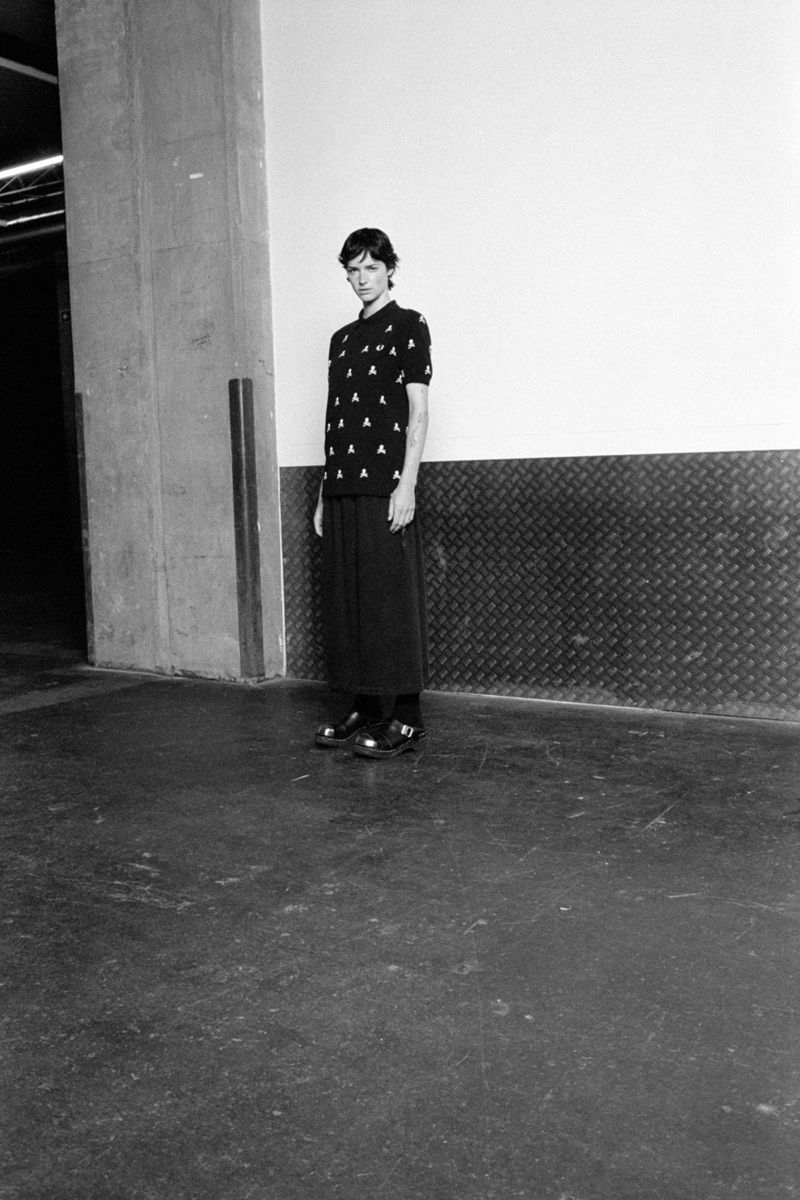Black and white full shot of a slender woman in a patterned black polo shirt and long black skirt standing indoors in a studio.