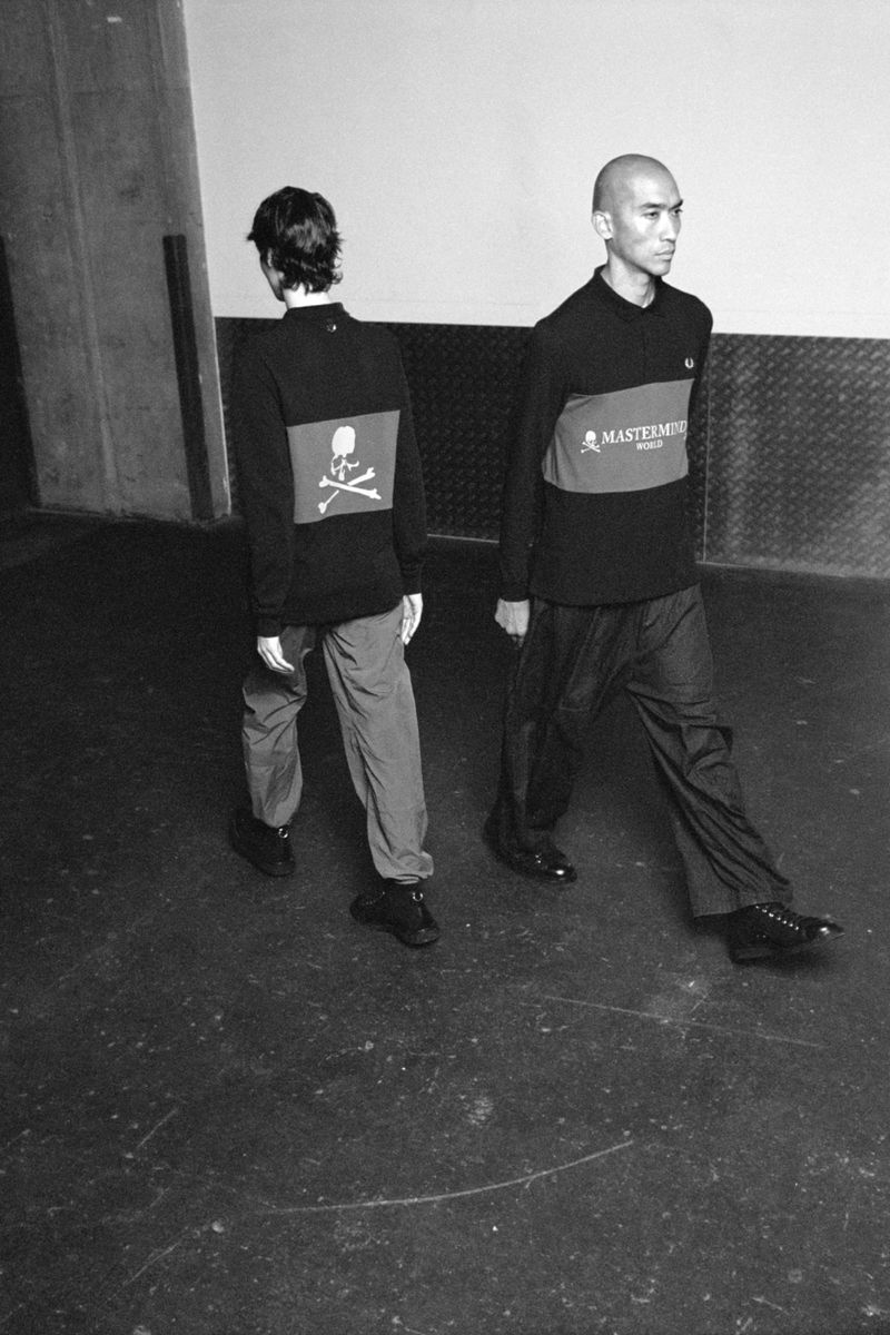Two men walk in long-sleeved shirts with skull logos and casual pants. Black and white image in an indoor setting, fashion-forward.