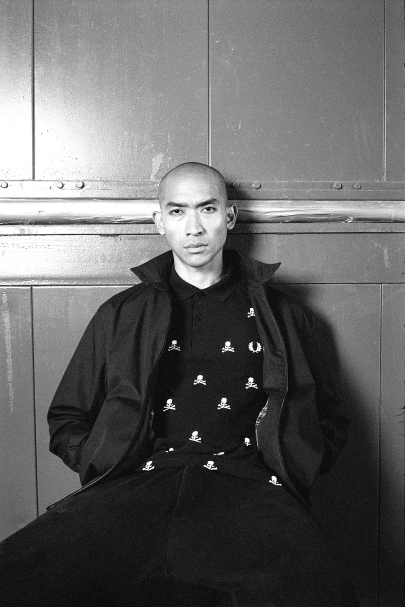 Monochrome portrait of a person with a shaved head in a black jacket and black polo shirt with skull and crossbones pattern against a metal background.