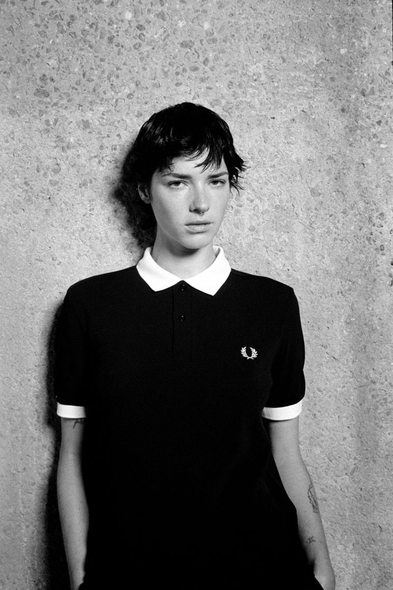 Monochrome portrait of a young adult woman in a black polo shirt with a white collar, posing in front of a textured wall.