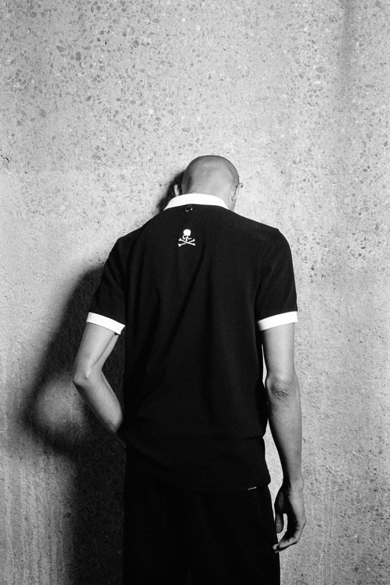 Model standing with back to camera wearing a black polo shirt with white trim against a textured concrete wall