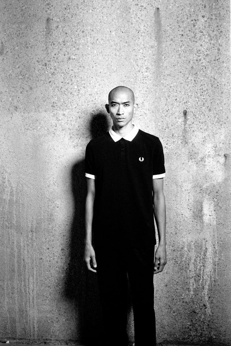 Monochrome portrait of a bald man in a dark polo shirt standing against a textured concrete wall, captured in stark, high-contrast lighting.