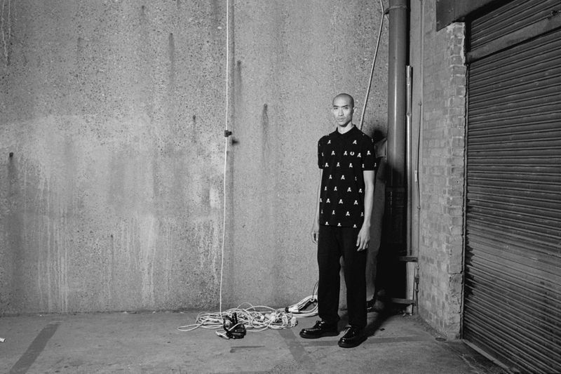 Monochrome portrait of a bald man in a patterned polo shirt standing in an industrial urban setting, with cables and roll-up door visible.