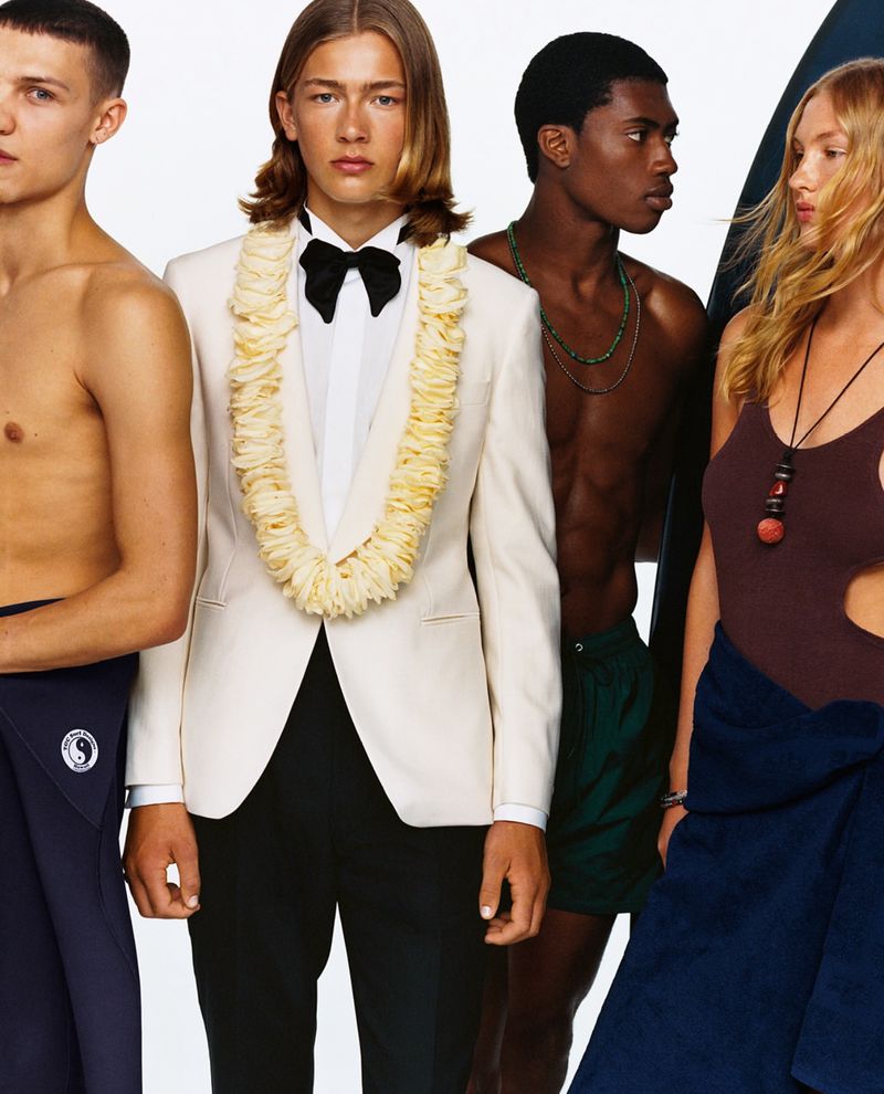 Four young adults are posing indoors. One man wears a white suit jacket and lei, while others wear casual attire, against a white backdrop.