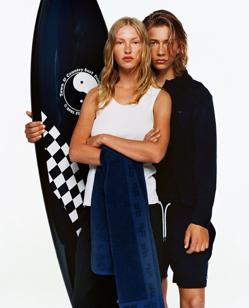 Two fashion models pose together in a studio with a surfboard and towel. The blonde woman wears a white tank top and blue towel.