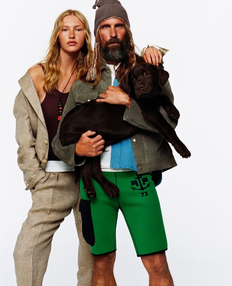 Editorial fashion shot of a stylish adult woman in beige suit with a man holding a dog, with green shorts and a beanie hat against a white backdrop.