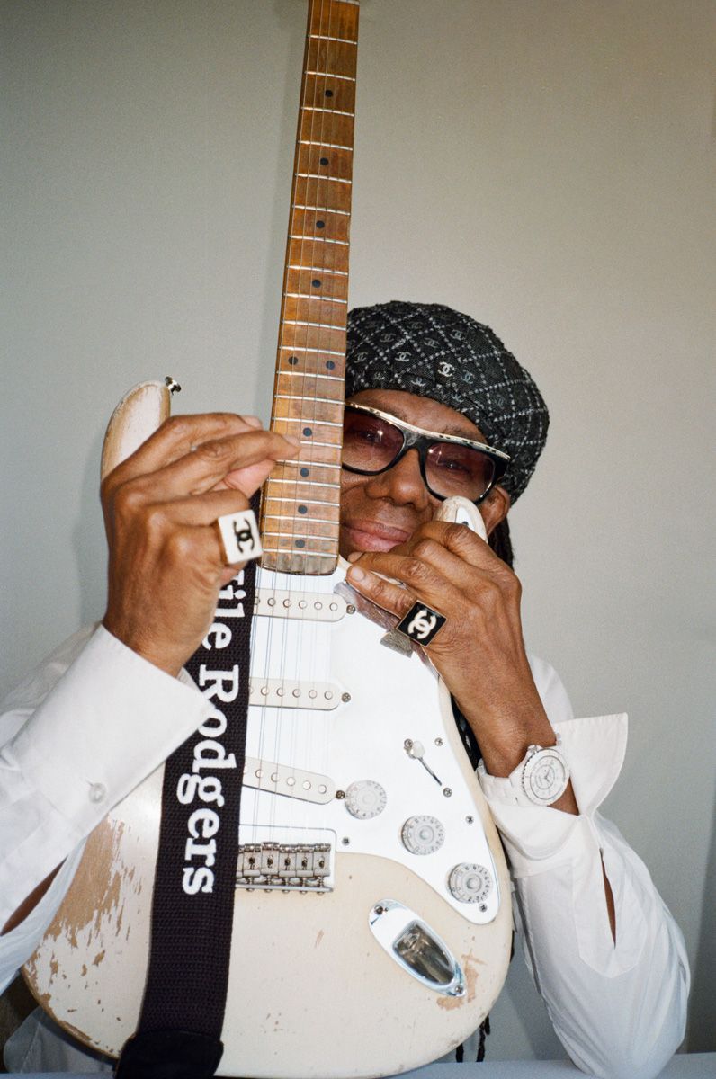 Portrait of Nile Rodgers with a white electric guitar, wearing black sunglasses, a patterned hat, rings and a white dress shirt.