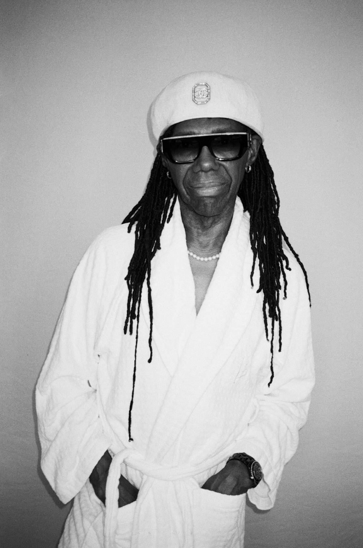 Black and white portrait of an adult male with dreadlocks, sunglasses, a white beret, and a white robe standing indoors.