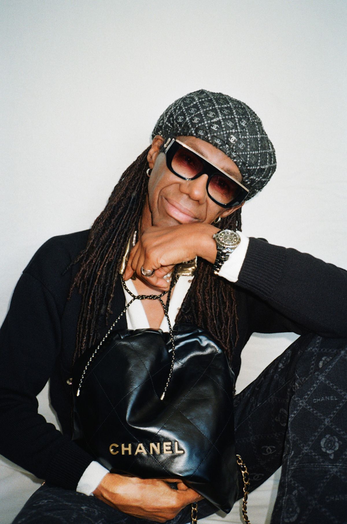 Stylish adult with dreadlocks poses indoors with a Chanel purse, sunglasses, and patterned beret. Wristwatch and patterned pants.