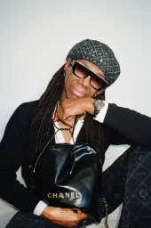Man with dreadlocks wearing oversized sunglasses, patterned beret, and black sweater holding a Chanel leather handbag.