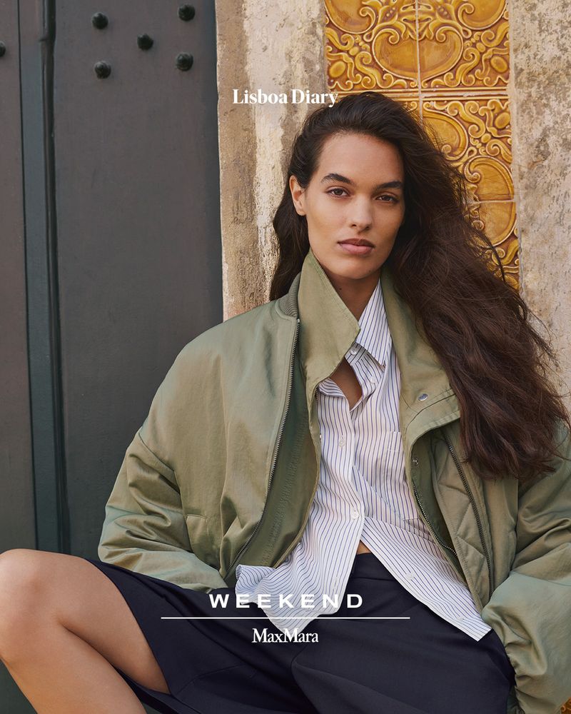 Fashion shot of a woman with long brown hair wearing an open olive-green jacket and striped shirt while sitting outside a stone building.