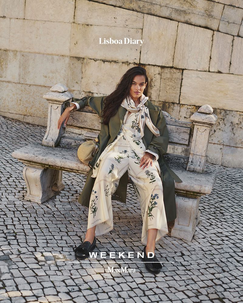 Model seated on a stone bench wearing an olive green coat over a patterned floral outfit for Weekend MaxMara