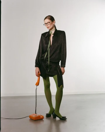 Fashionable woman in black blouse, skirt, and green tights, holding an orange landline telephone against a neutral backdrop in a studio.