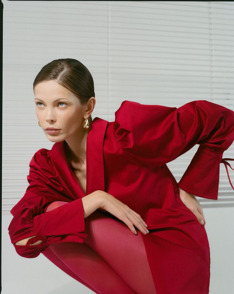 Fashion portrait of a woman in a red satin blouse and red leggings in a studio setting posing against a white wall with blinds.