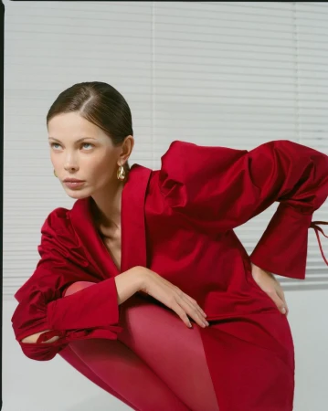 Fashion portrait of a woman in a red satin blouse and red leggings in a studio setting posing against a white wall with blinds.