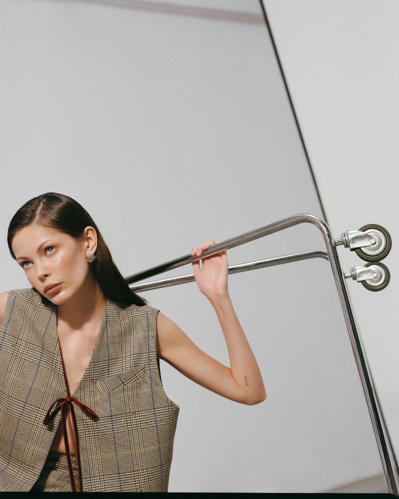 Brunette woman poses with a metal clothes rack in a fashion shot, wearing a brown plaid vest with a ribbon closure in a photo studio.