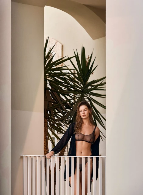 A young adult female model poses in lingerie and a sheer black robe on a balcony in front of a yucca plant inside a white building.