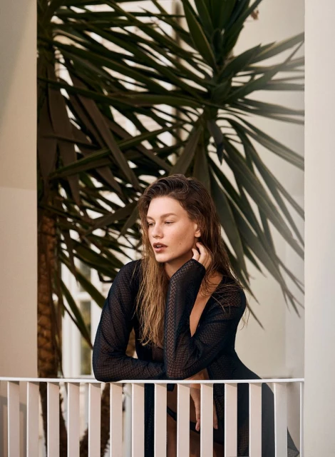 Portrait of an adult woman wearing a sheer black top, leaning on a white railing, with a palm plant and interior view.
