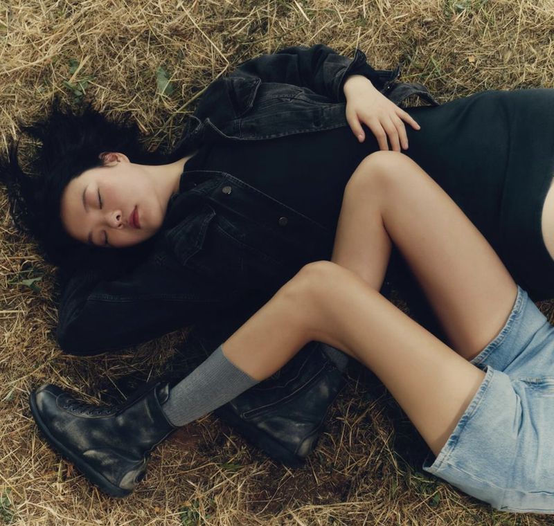 Two young women are lying on the grass. One is wearing a black denim jacket and the other is wearing a blue denim skirt and black boots.