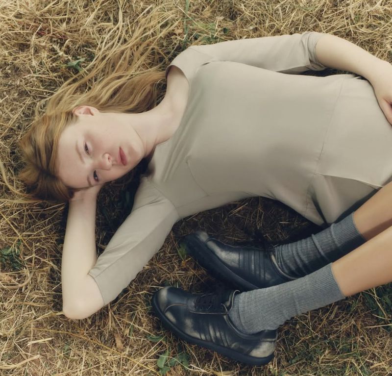 Woman with red hair reclines on a grassy surface in a light tan dress and black shoes, posing in a natural outdoor setting.