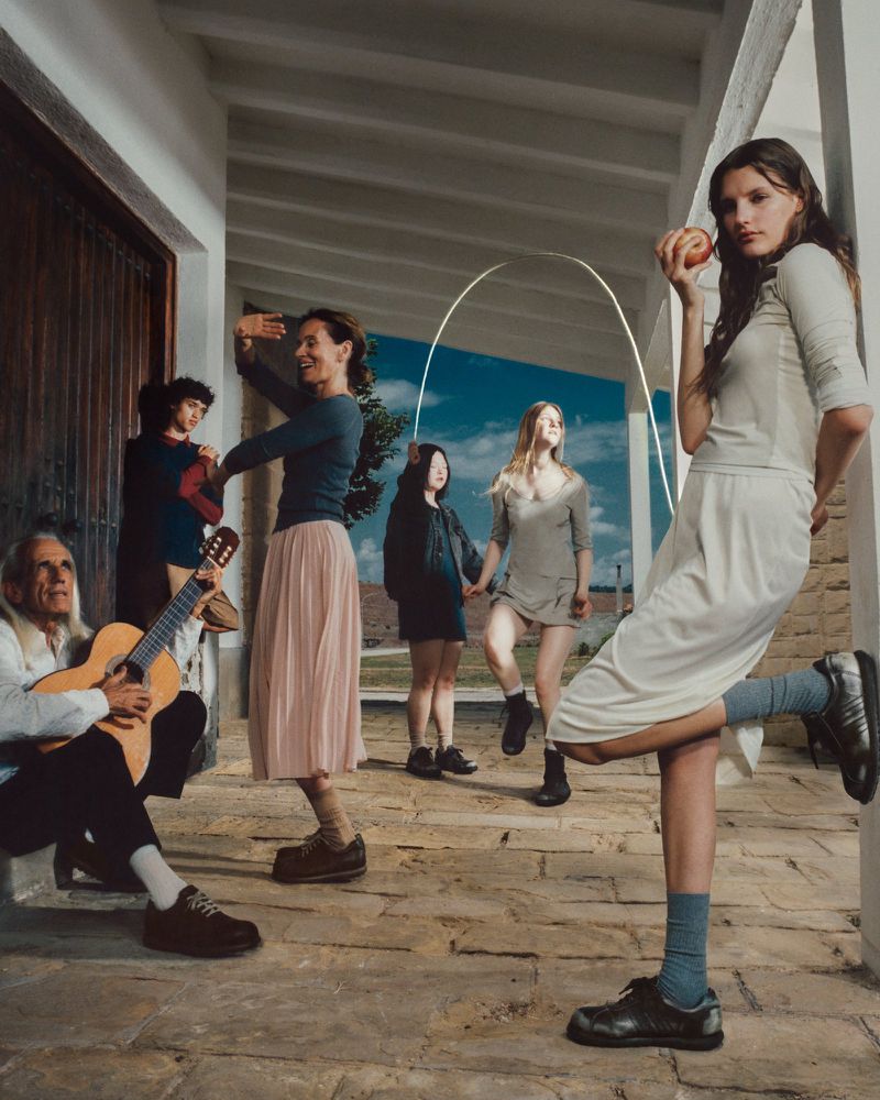Group of people gathered outdoors on a veranda, including a woman holding an apple and other people dancing or playing a guitar.