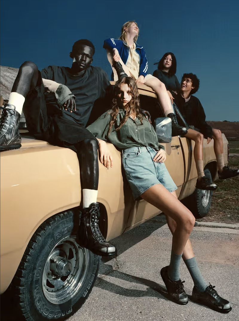 Group of five people posing on an old, yellow pickup truck against a blue sky and arid landscape. They are dressed in casual clothing.