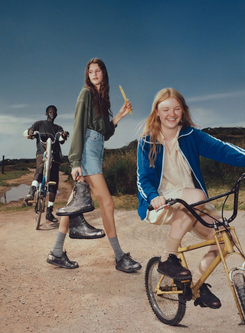 Three young people with bicycles on a dirt road under a blue sky; one holds black work boots; lifestyle fashion editorial.