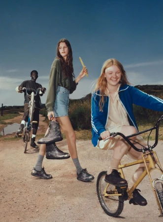 Three young people with bicycles on a dirt road under a blue sky; one holds black work boots; lifestyle fashion editorial.