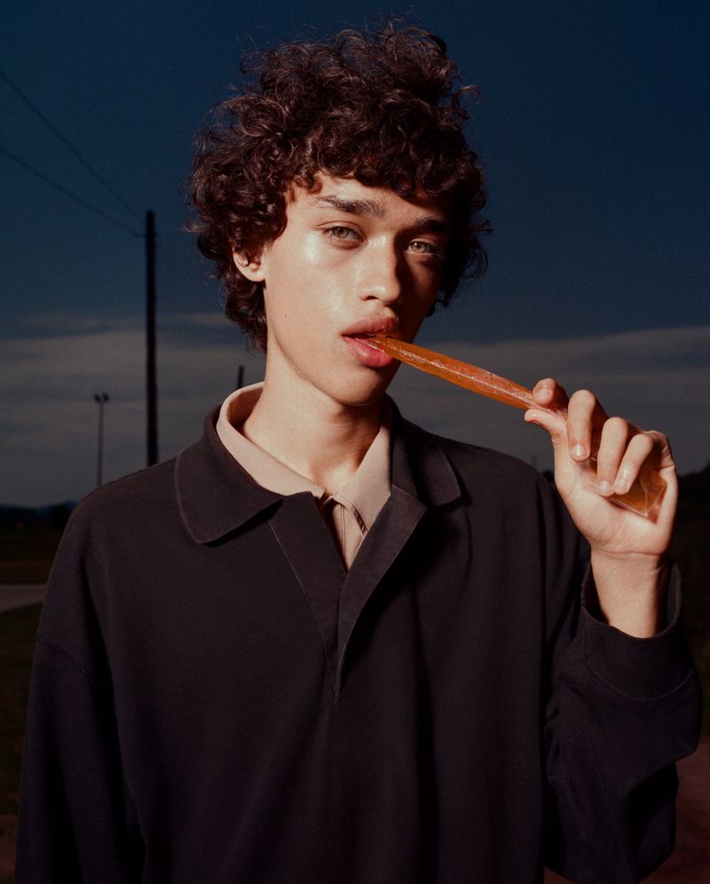 Portrait of a young adult male model with curly hair wearing a black polo, holding a translucent orange snack, outdoor setting, medium shot.