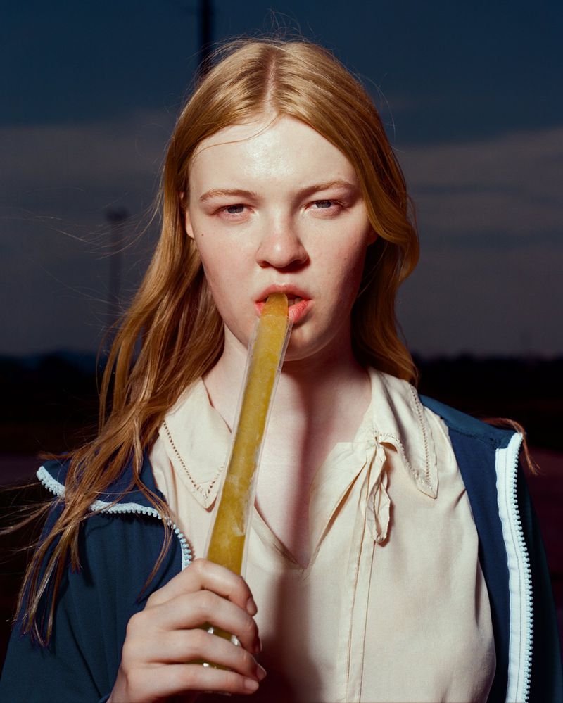 A young woman with red hair eats a yellow popsicle in an outdoor dusk setting, wearing a beige top and blue jacket.