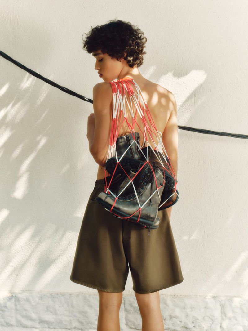 Young adult with curly hair carries boxing gloves in a string bag over his shoulder in a fashion shot against a white wall.
