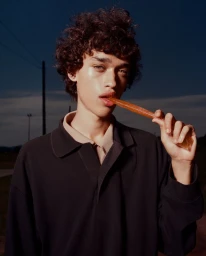 A young male model with dark curly hair wearing a dark polo shirt holding a snack against an evening sky background for Camper