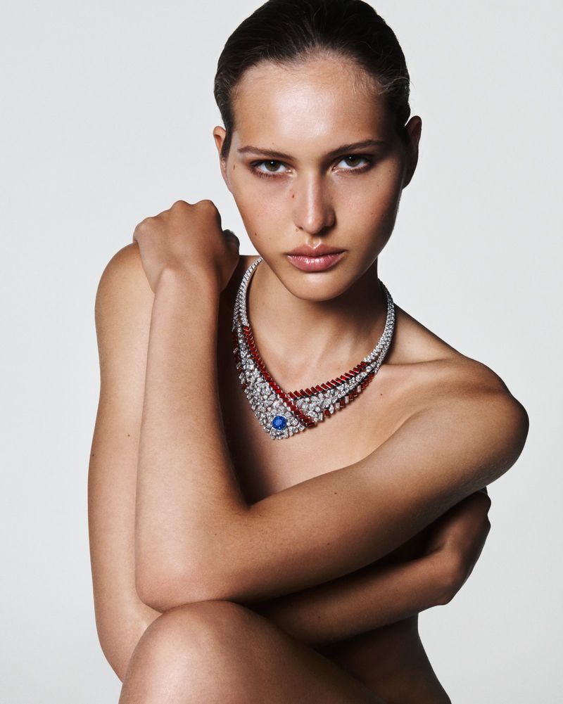 Elegant woman poses in a close-up shot wearing a statement necklace with ruby, diamond, and sapphire gemstones against a white backdrop.