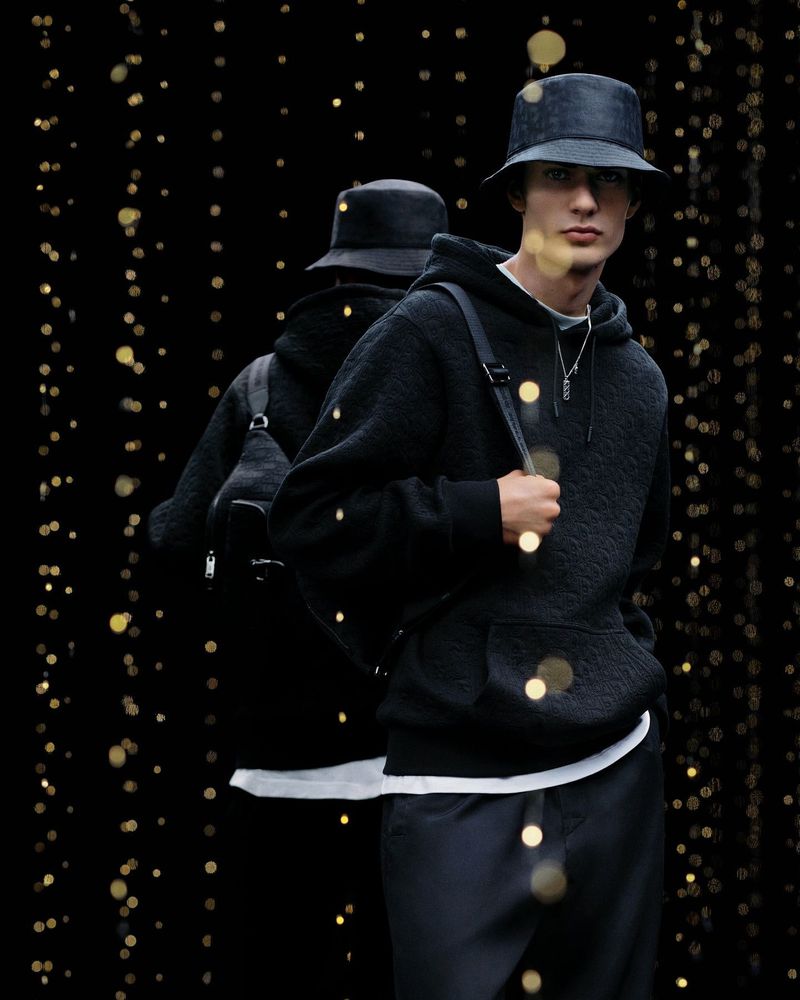 Young man in dark textured hoodie, pants, and bucket hat poses against a black backdrop with bokeh lights and reflection.