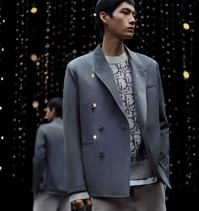 An adult male fashion model poses in a grey blazer and patterned sweater against a bokeh background, creating a stylish studio portrait.