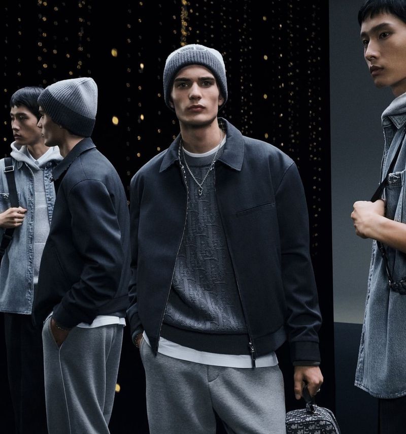 Three adult male models in gray knit beanies, sweaters, jackets, and sweatpants stand in a studio with a dark, speckled background.