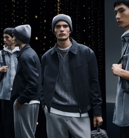Three adult male models in gray knit beanies, sweaters, jackets, and sweatpants stand in a studio with a dark, speckled background.