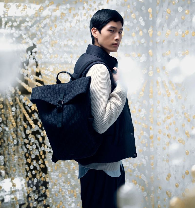 Male model showcases a Dior backpack in an eye-level shot, wearing a sweater and vest against a backdrop of white and gold bokeh.