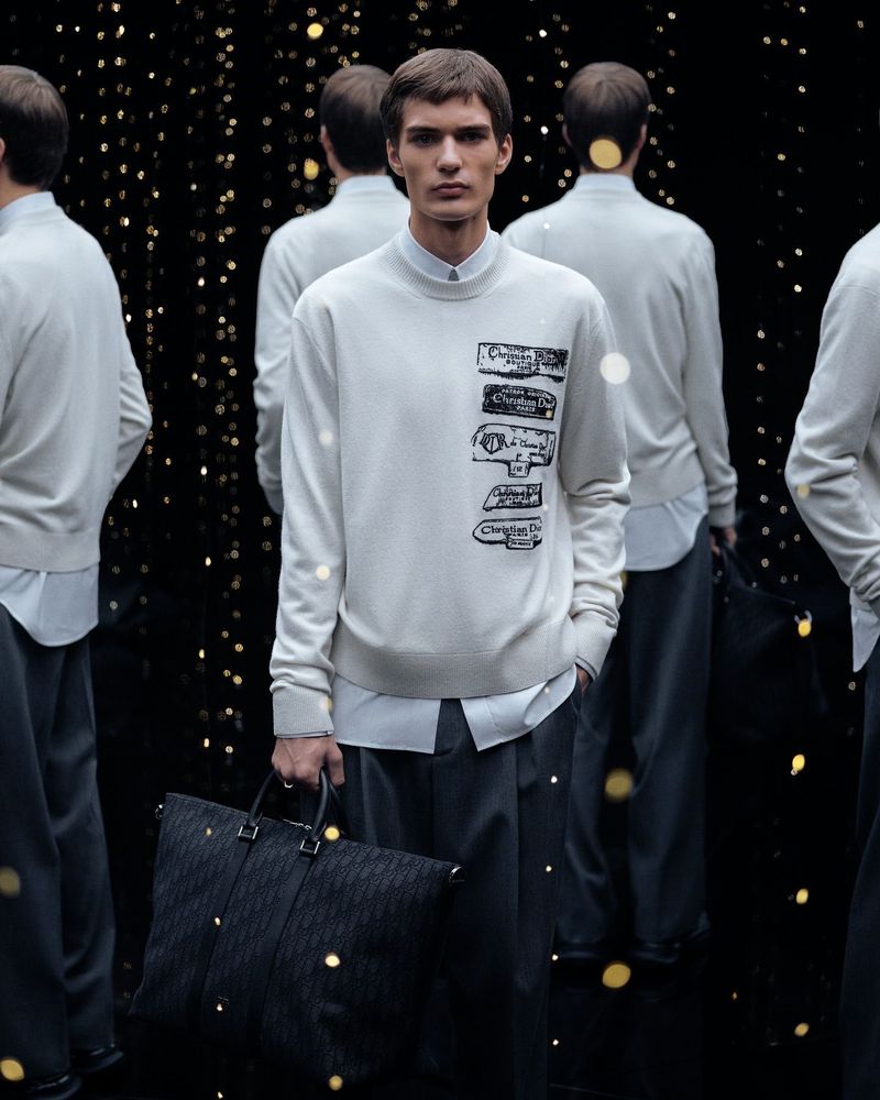 Male fashion model wearing a white sweater with logos holding a black tote bag surrounded by other figures and bokeh lights.