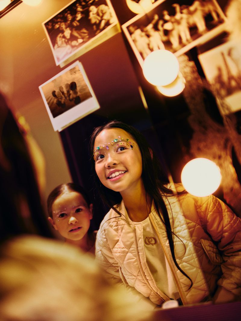 Two young girls, one Asian, with decorative face jewels and light jackets pose indoors with photo decorations and warm, glowing light bulbs.