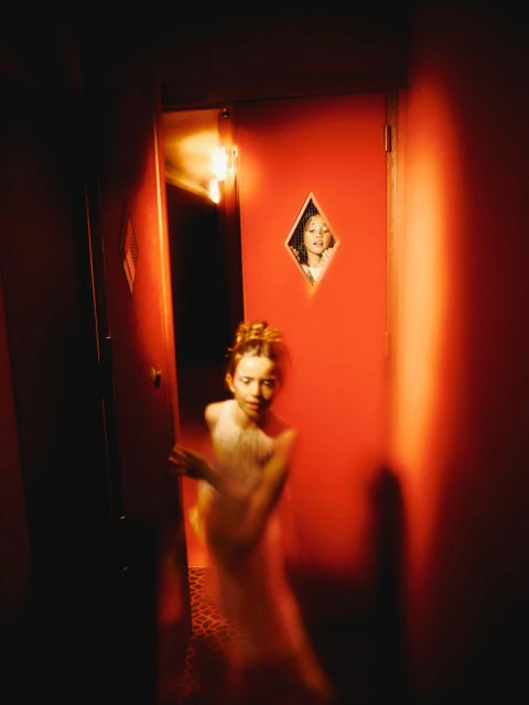 A girl stands in a doorway in a dimly lit red room while another child looks through a diamond shaped window in the red door.