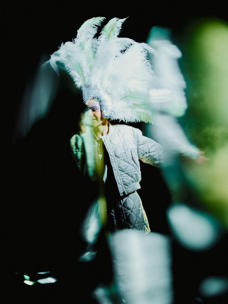 Woman in a quilted jacket and ornate feathered headpiece poses in the dark, illuminated by blue and green lighting.