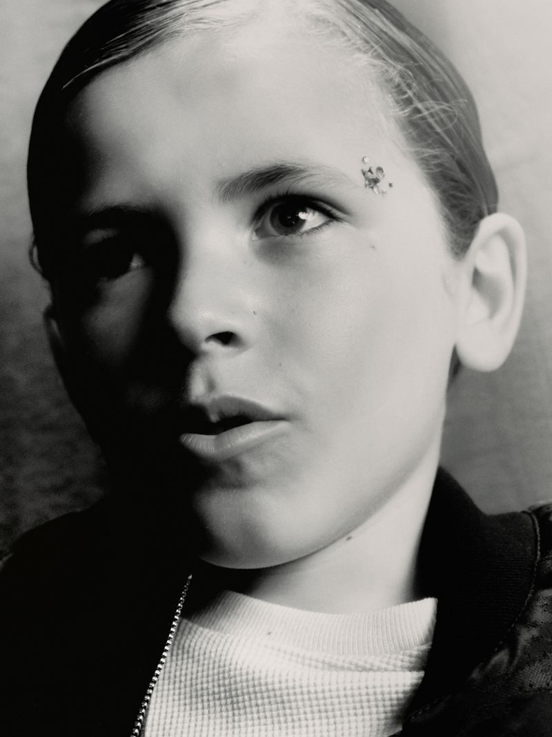Black and white portrait of a young boy with a spot on his forehead wearing a waffle-knit shirt and a bomber jacket.