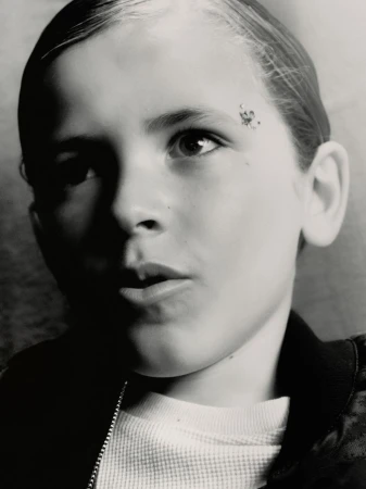 Black and white portrait of a young boy with a spot on his forehead wearing a waffle-knit shirt and a bomber jacket.