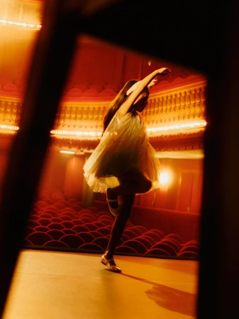 Female dancer in mid-air on a stage of an old theater with red velvet seats and ornate gold details, creating a vintage performance scene.
