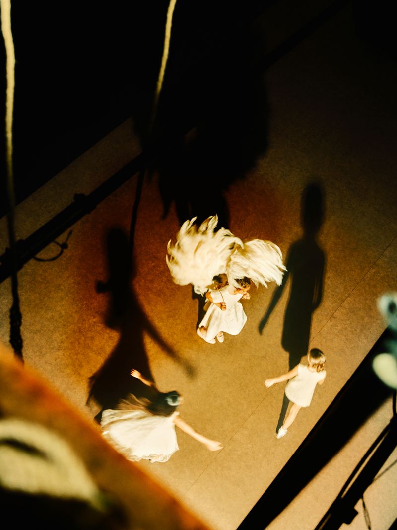 Aerial shot features four girls in white dresses on a tan surface, with dramatic shadows and glimpses of rigging, creating an artistic, ethereal atmosphere.